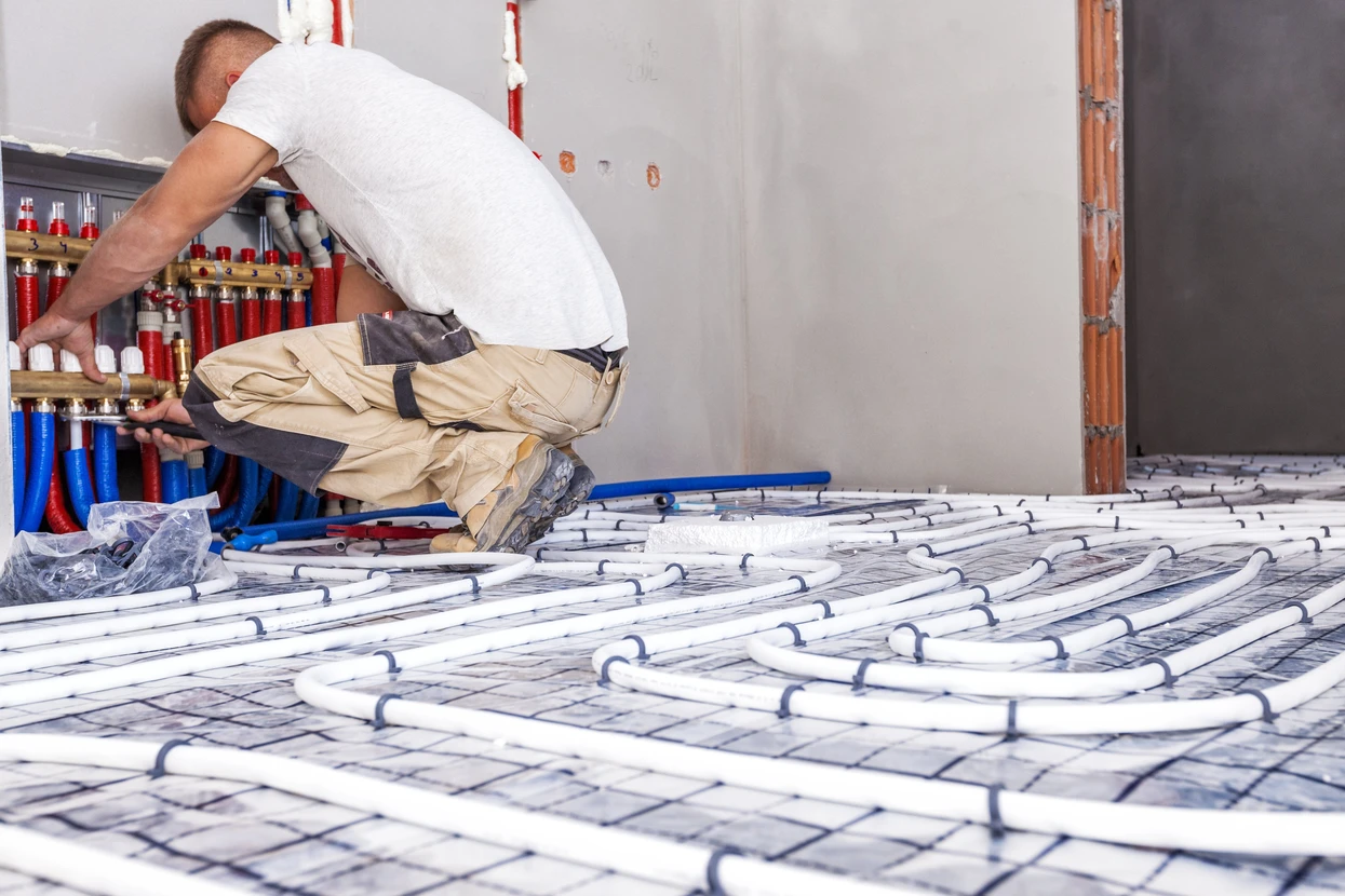 Installateur en combinaison beige travaille sur système de plancher chauffant hydraulique, connectant tuyaux PEX au collecteur-répartiteur avec vannes rouges et bleues